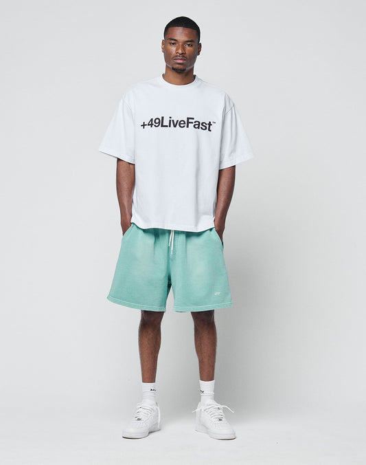 A man faces forward against a plain light background, wearing the LIVE FAST DIE YOUNG Basic +49 Tee (100% Baumwolle) with front print, mint green shorts, white socks, and sneakers. His hands are in his pockets.