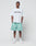 A man faces forward against a plain light background, wearing the LIVE FAST DIE YOUNG Basic +49 Tee (100% Baumwolle) with front print, mint green shorts, white socks, and sneakers. His hands are in his pockets.