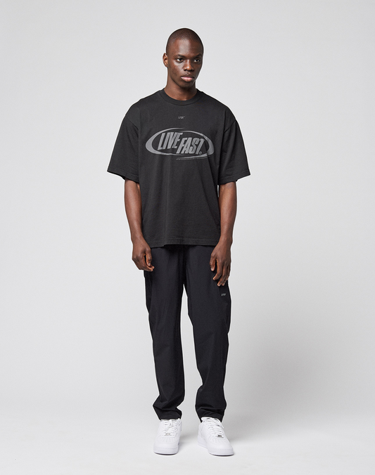 A man stands facing forward against a plain background, wearing a black T-shirt with LIVE FAST and a reflective logo print, LFDY Performance Pants by LIVE FAST DIE YOUNG, and white sneakers. His arms rest naturally at his sides.