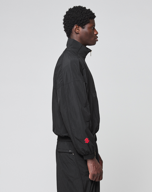 A person with short curly hair stands in profile against a plain background, wearing the LIVE FAST DIE YOUNG LFDY Dealership Trackjacket—black with a Stehkragen and a small red embroidered detail on the sleeve—paired with matching black pants.