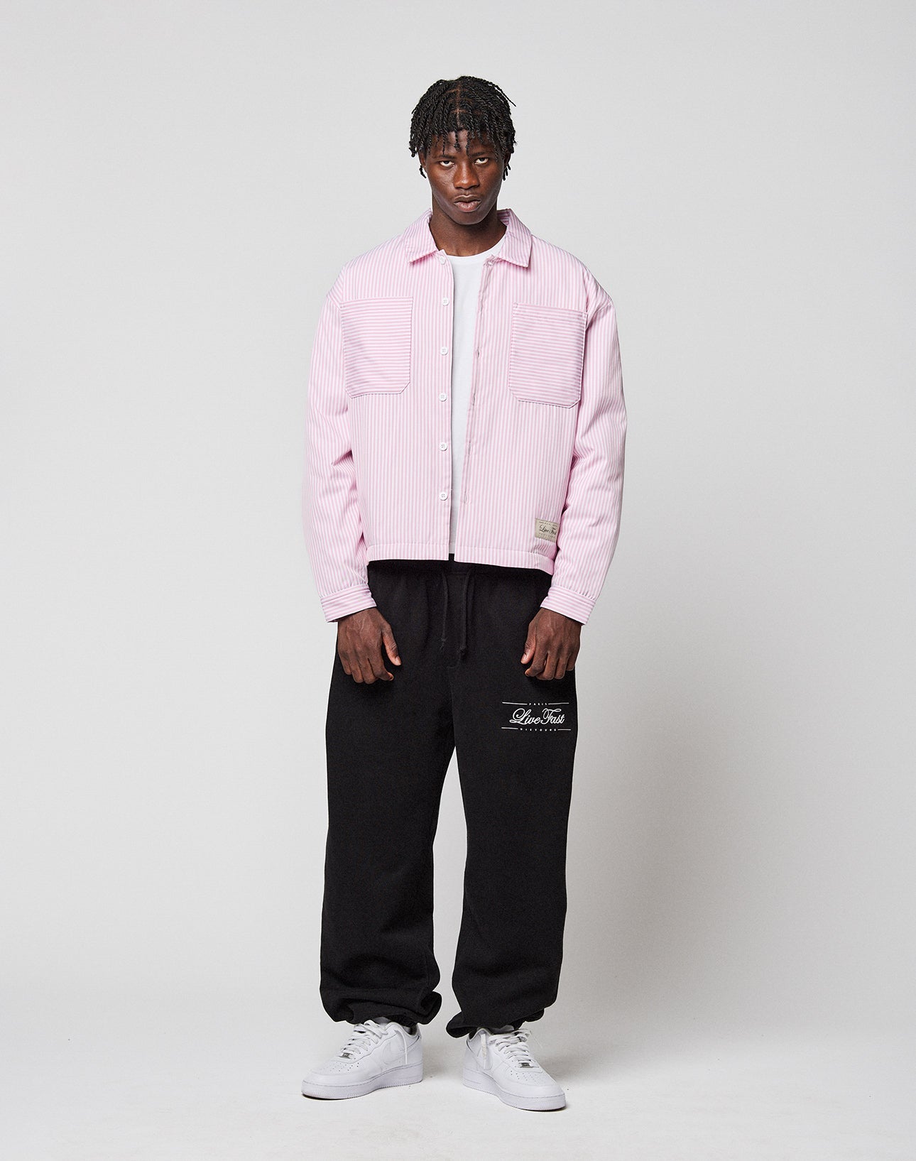 A man stands against a white background wearing the LIVE FAST DIE YOUNG LFDY Striped Padded Shirt over a white shirt, black sweatpants with white text, and white sneakers. He faces forward with a neutral expression.