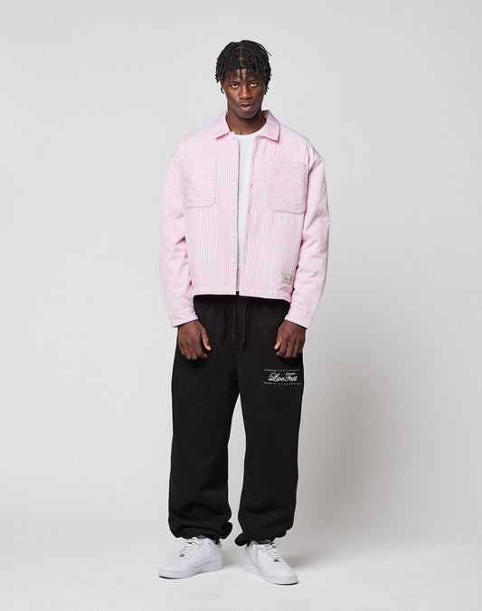 A man stands against a white background wearing the LIVE FAST DIE YOUNG LFDY Striped Padded Shirt over a white shirt, black sweatpants with white text, and white sneakers. He faces forward with a neutral expression.