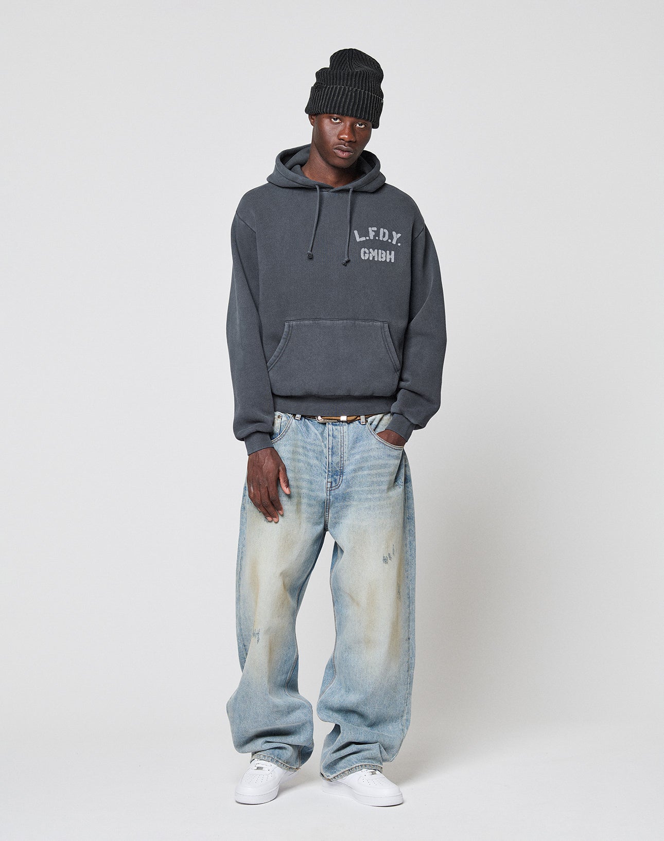 A person in a black beanie, LIVE FAST DIE YOUNG LFDY GmbH Hooded dark gray hoodie with Kapuze and text, baggy light blue oversize jeans, and white sneakers stands against a plain light background.