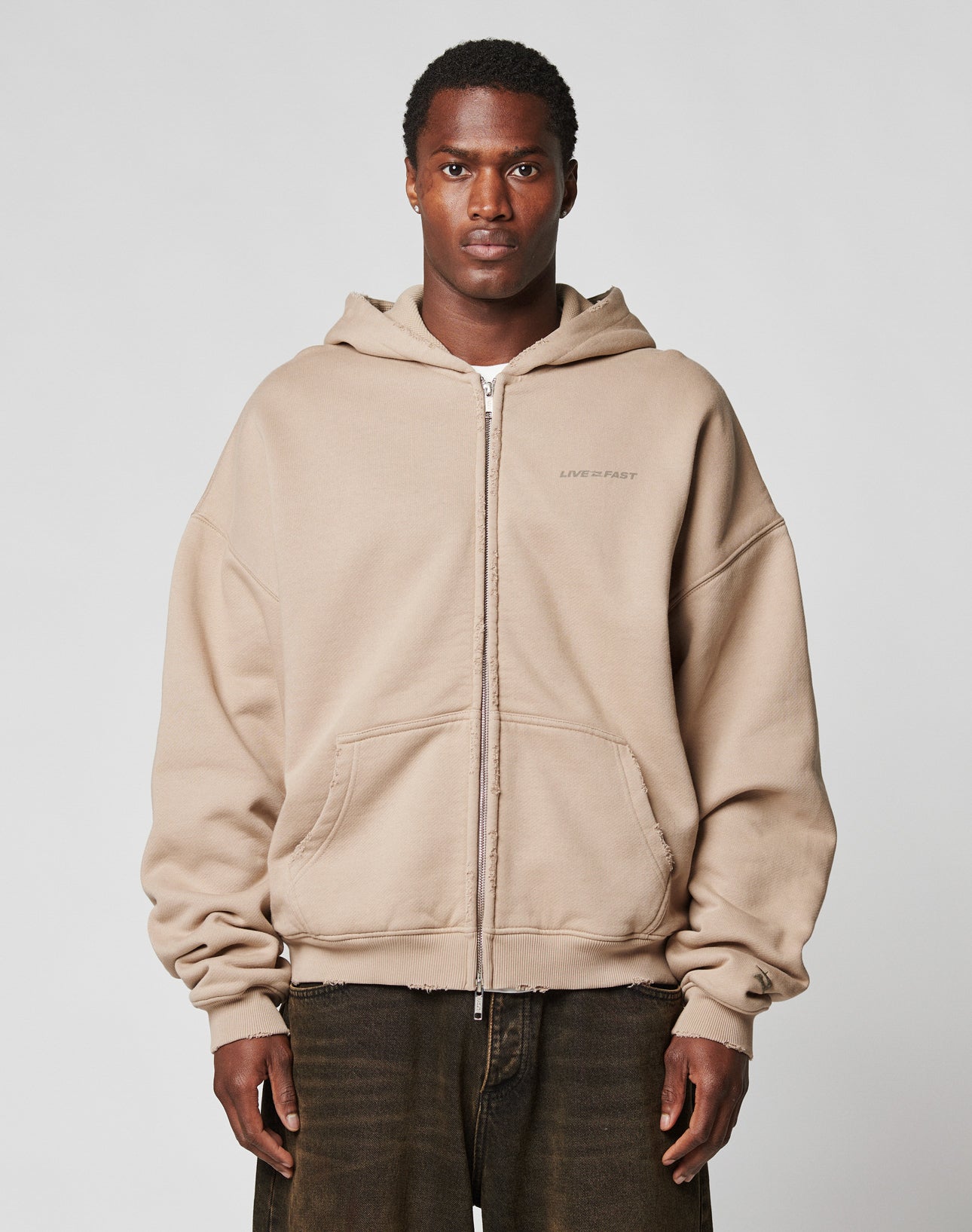 A person stands facing forward in a beige LIVE FAST DIE YOUNG LFDY Double Hooded Zip with subtle waffle lining and front pockets, paired with dark brown pants against a plain, light background.