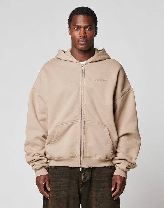 A person stands facing forward in a beige LIVE FAST DIE YOUNG LFDY Double Hooded Zip with subtle waffle lining and front pockets, paired with dark brown pants against a plain, light background.