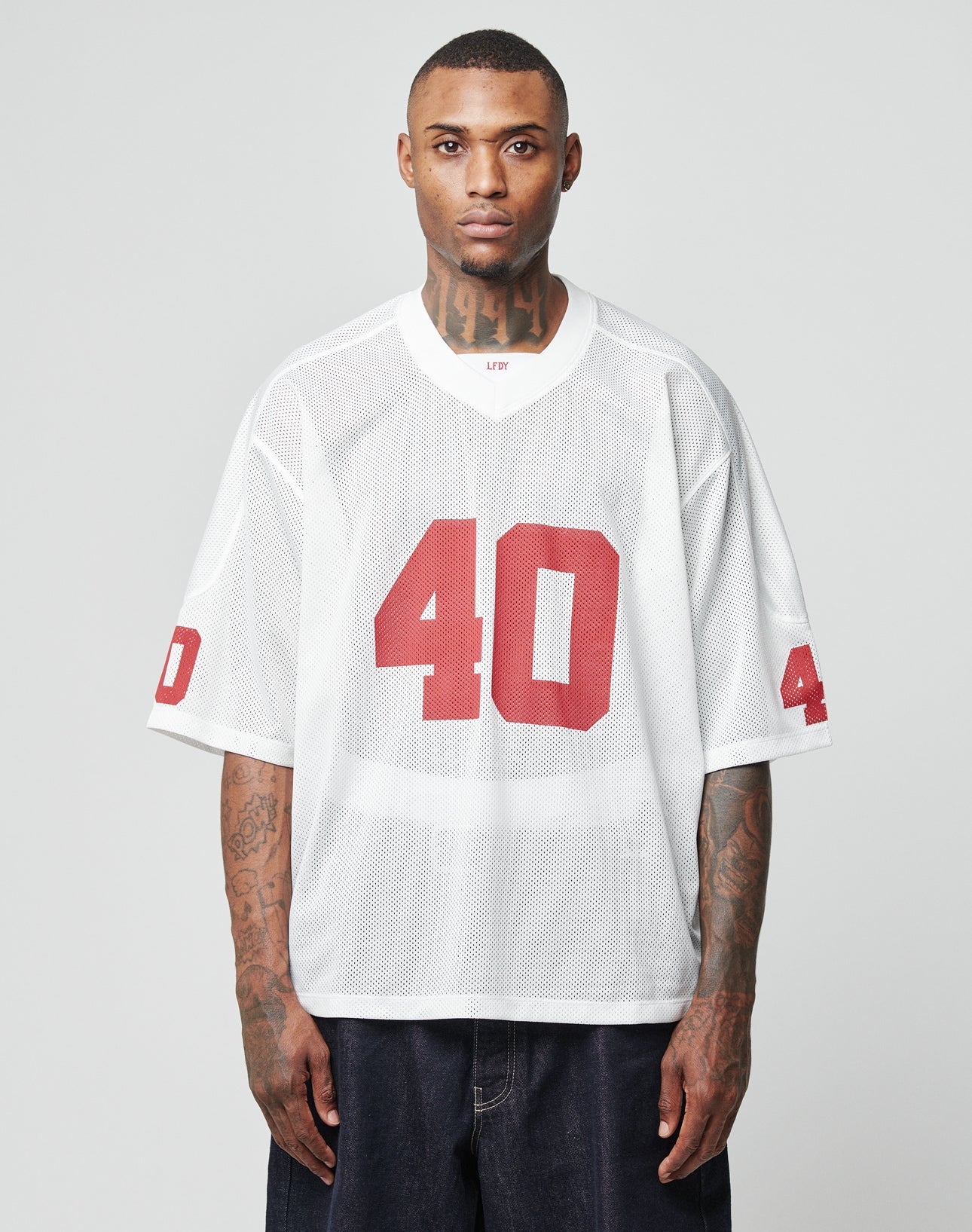 A tattooed man wears the LFDY Mesh Jersey by LIVE FAST DIE YOUNG—an oversized white mesh polyester top with a V-neck and red number 40 on front and sleeves—facing forward against a plain light gray background. | model