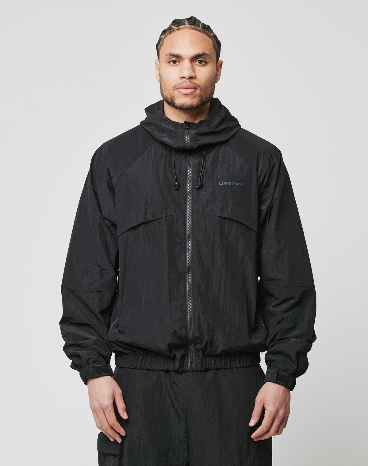 A person stands facing forward, wearing the LIVE FAST DIE YOUNG Live Fast Nylon Jacket in black with seitliche Eingrifftaschen and an oversized fit, paired with matching pants, hands relaxed by their sides against a plain light grey background. | model