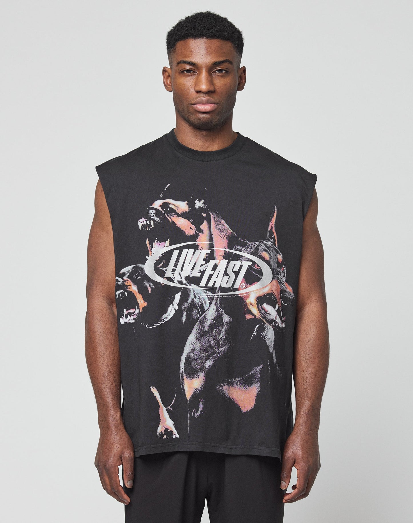 A man faces forward in a LFDY Canine Muscle Shirt by LIVE FAST DIE YOUNG—an oversize, sleeveless black top with snarling dogs and “LIVE FAST” in silver lettering. The background is plain and light-colored.