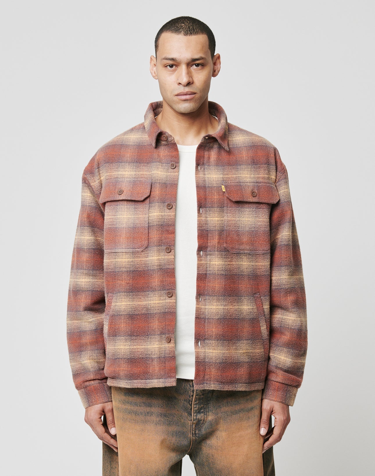 A man faces forward wearing the LIVE FAST DIE YOUNG Vintage Padded Flannel in red and beige plaid over a white shirt and brown pants, set against a plain light gray background. | model