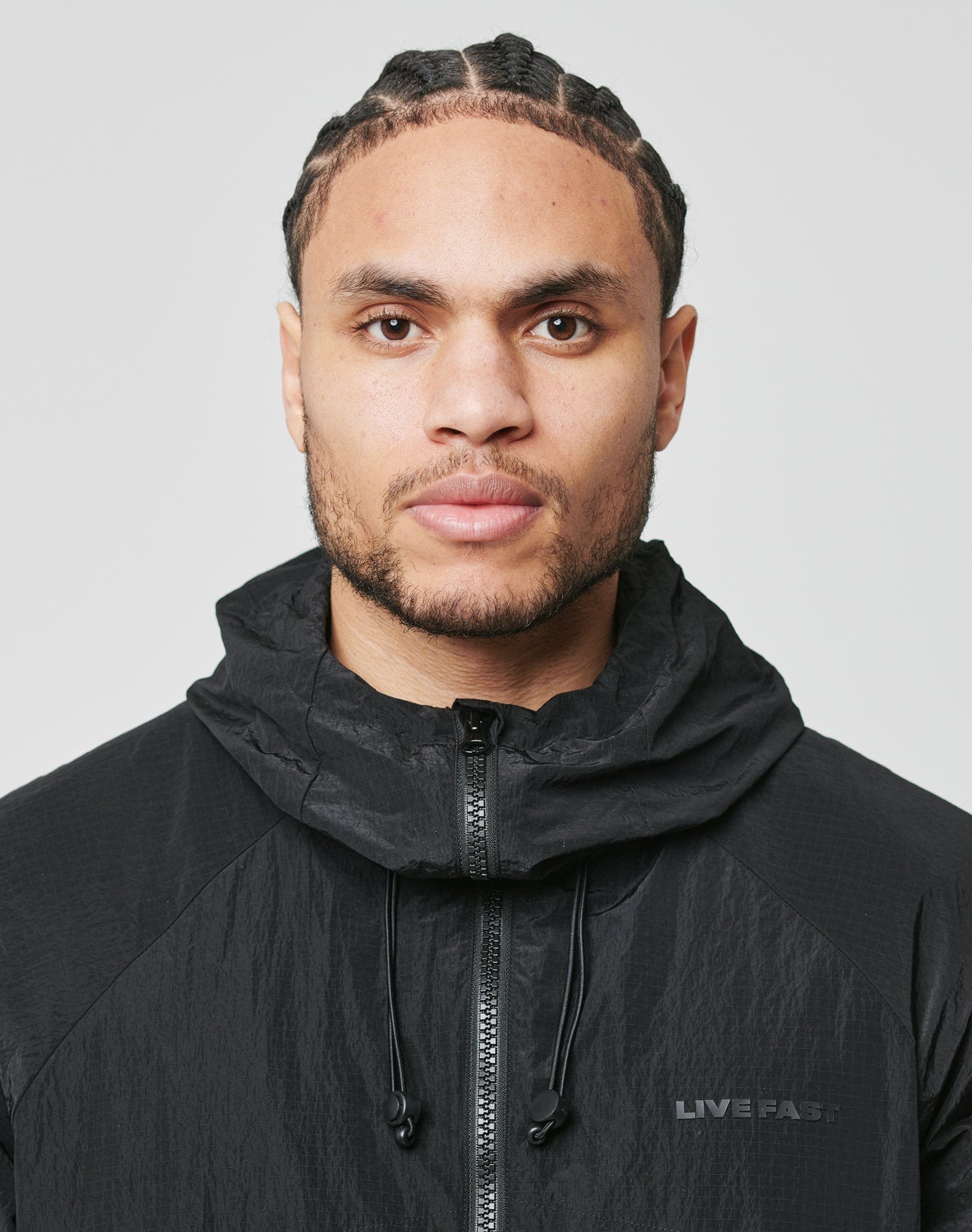 A man with braided hair and a trimmed beard faces forward against a plain background, wearing the LIVE FAST DIE YOUNG Live Fast Nylon Jacket—an oversized black zip-up with LIVE FAST in silicone print.