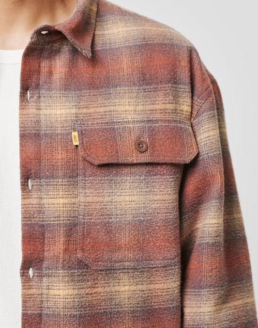 A person wears the LIVE FAST DIE YOUNG Vintage Padded Flannel in brown, rust, and beige plaid over a white textured shirt, highlighting its buttoned chest pocket and part of the collar.
