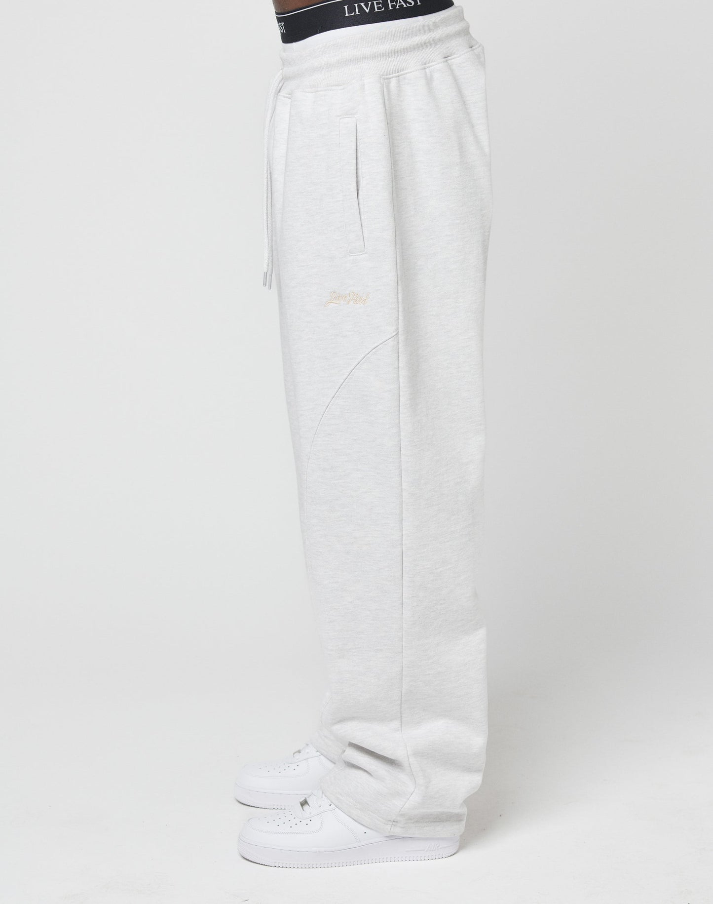 A person wearing the LIVE FAST DIE YOUNG Basic Wide Jogger in light gray with an elastic waistband and wide fit, along with white sneakers and a black LIVEFAST waistband, stands sideways against a plain white background.