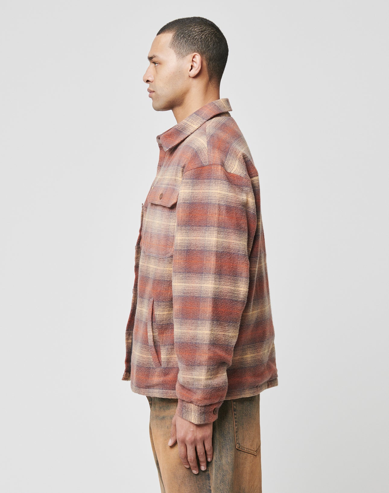 A man stands in profile against a plain background, wearing the LIVE FAST DIE YOUNG Vintage Padded Flannel—an oversize fit brown, beige, and red plaid shirt jacket with front pockets—paired with tan pants.