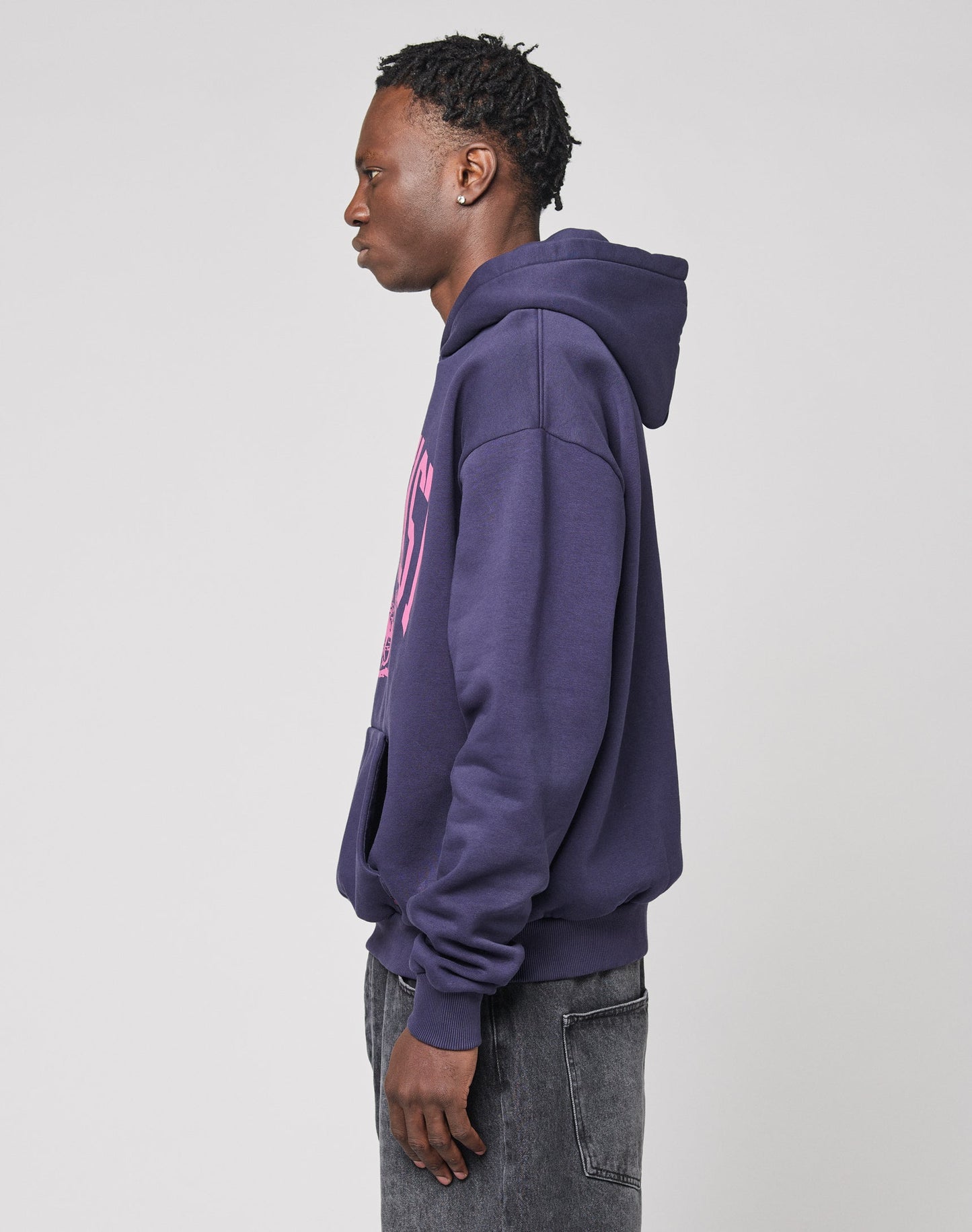 A young man stands in profile against a plain background, wearing the oversized Live Fast Stallion Hooded by LIVE FAST DIE YOUNG with a pink front print and faded black jeans.