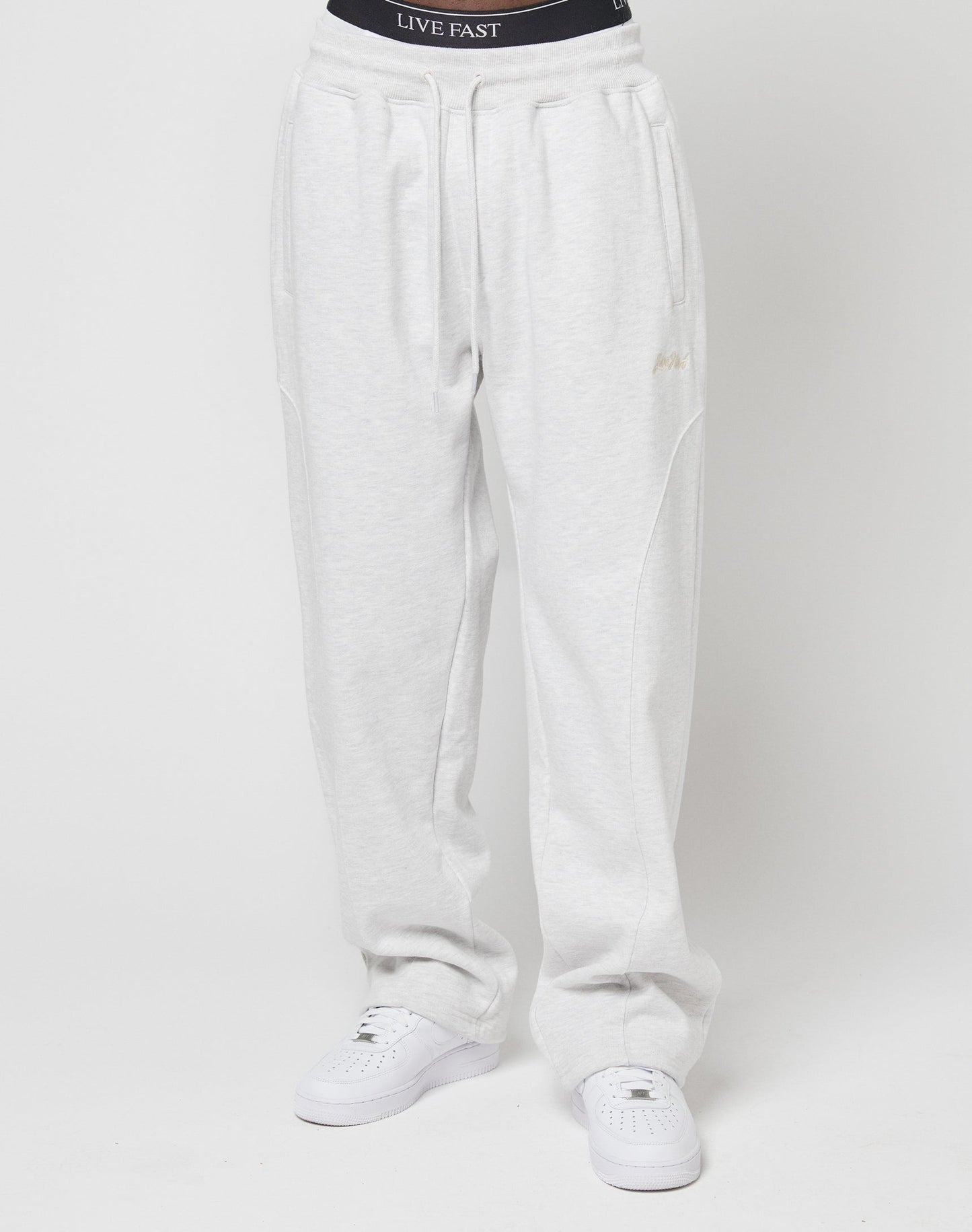 A person wears the Basic Wide Jogger by LIVE FAST DIE YOUNG in light gray with an elastic waistband that reads LIVE FAST, paired with white sneakers. The photo is cropped at the waist, so their upper body and head arent visible.