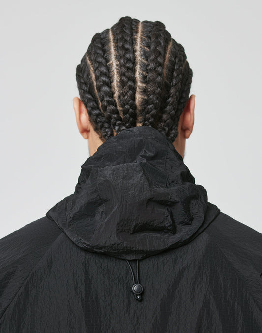 A person with neat cornrows is shown from behind, wearing the LIVE FAST DIE YOUNG Live Fast Nylon Jacket in black with textured fabric and side pockets, hood down, against a plain light background.