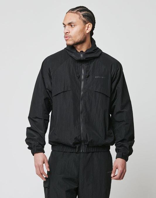 A man with braided hair wears the LIVE FAST DIE YOUNG Live Fast Nylon Jacket, an oversized black zip-up windbreaker with side pockets, and matching pants. He stands against a light gray background, looking left with one hand in his pocket.