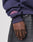 A close-up of a hand at the side, wearing the LIVE FAST DIE YOUNG Live Fast Stallion Hooded in dark purple with pink embroidered text and crossed swords on the sleeve cuff, paired with dark gray jeans.