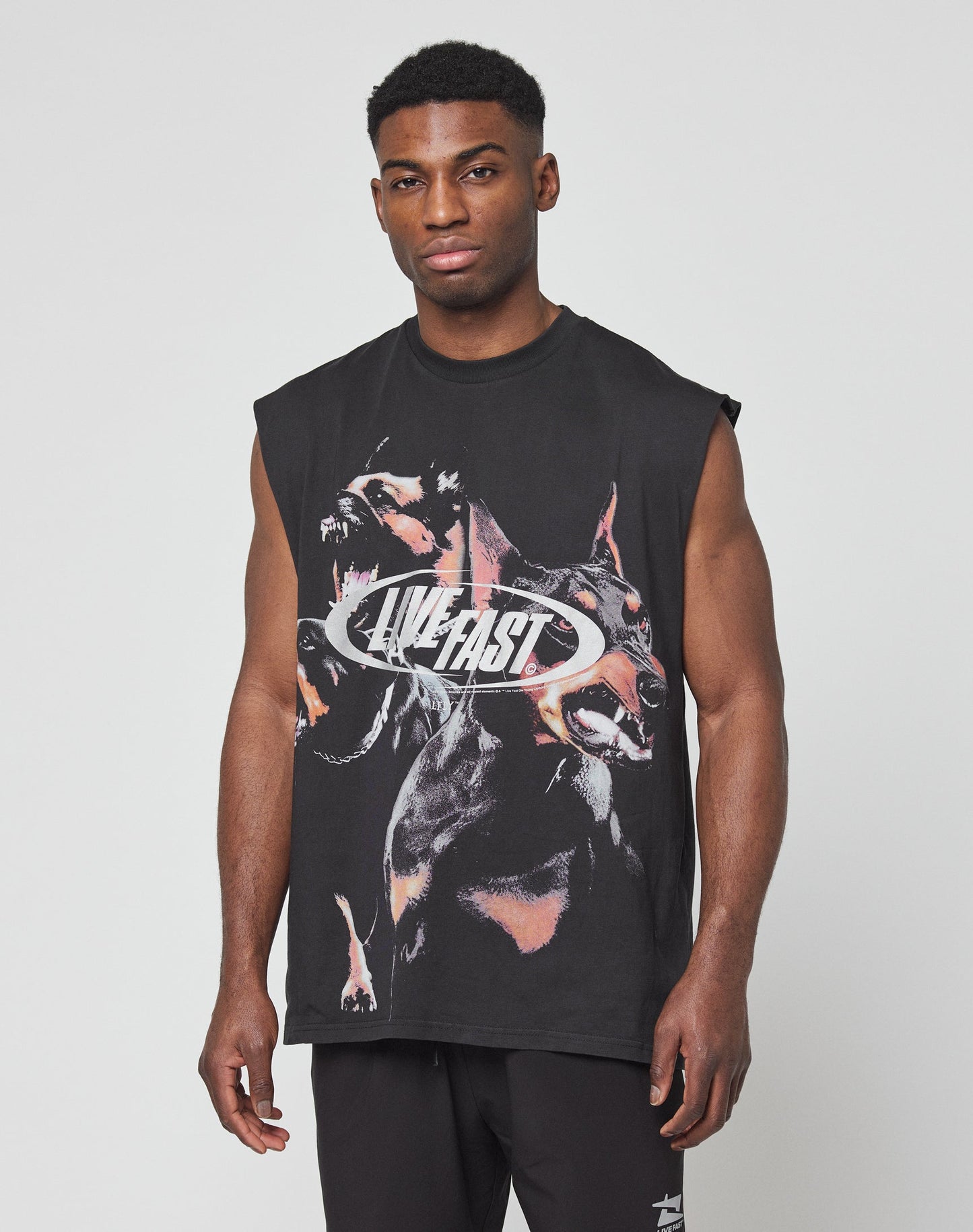 A man wears the LFDY Canine Muscle Shirt by LIVE FAST DIE YOUNG—a sleeveless black tee with two growling dogs and the silver LIVE FAST lettering on the front, made from 100% cotton—standing against a plain light background.