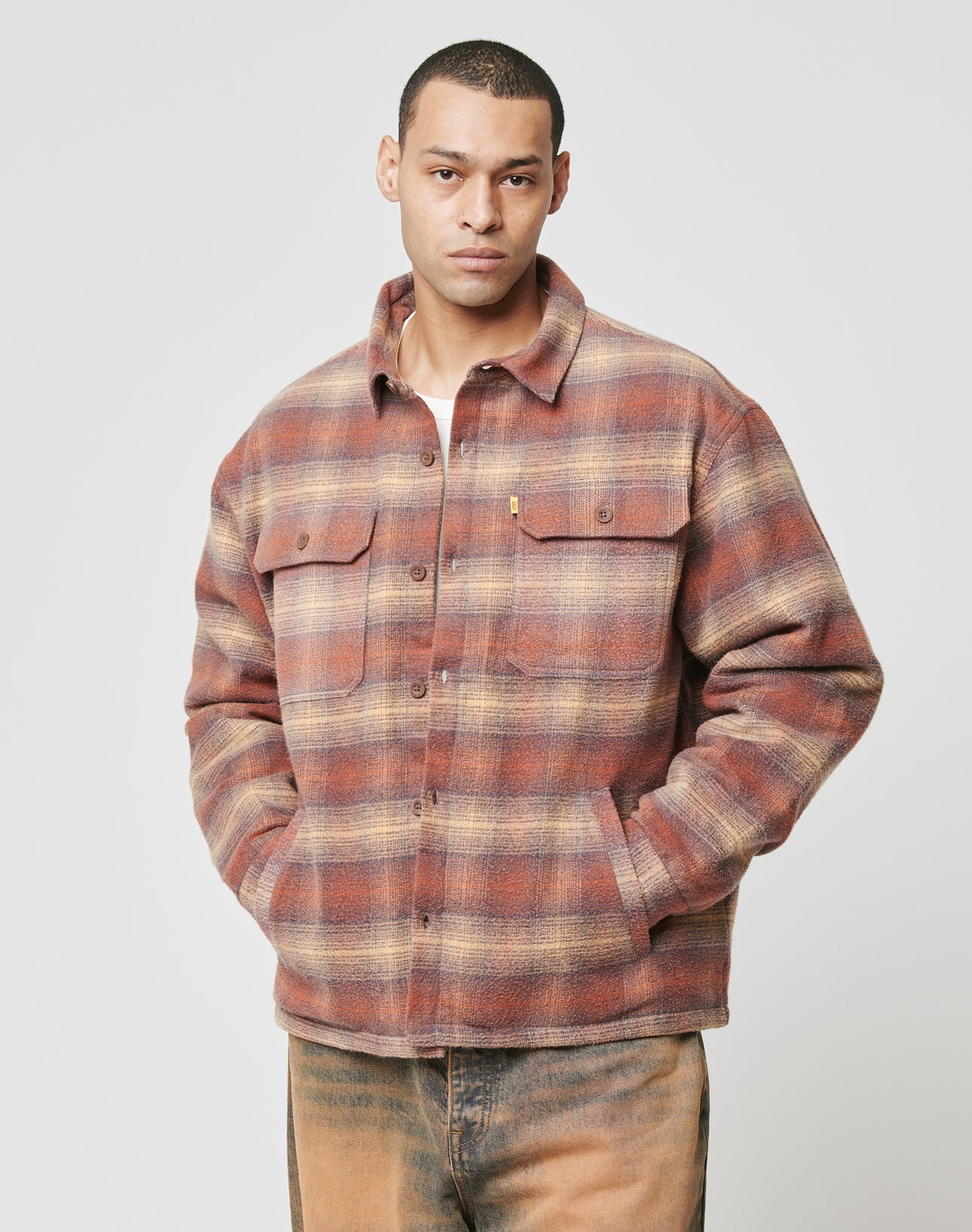 A man stands facing forward with hands in pockets, wearing the LIVE FAST DIE YOUNG Vintage Padded Flannel in brown and orange plaid and tan pants against a plain light background.