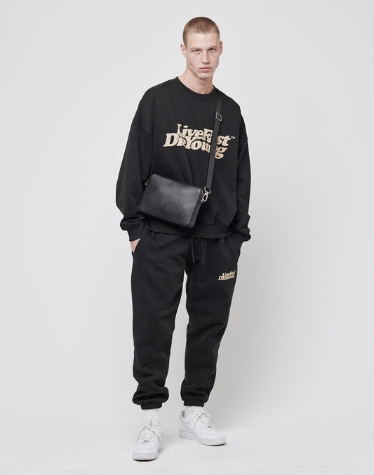 A young man stands against a white background wearing a black sweatshirt with beige text, matching sweatpants, white sneakers, and the LIVE FAST DIE YOUNG Airliner Bag in logo-embossed Rindsleder. He looks at the camera with a neutral expression.