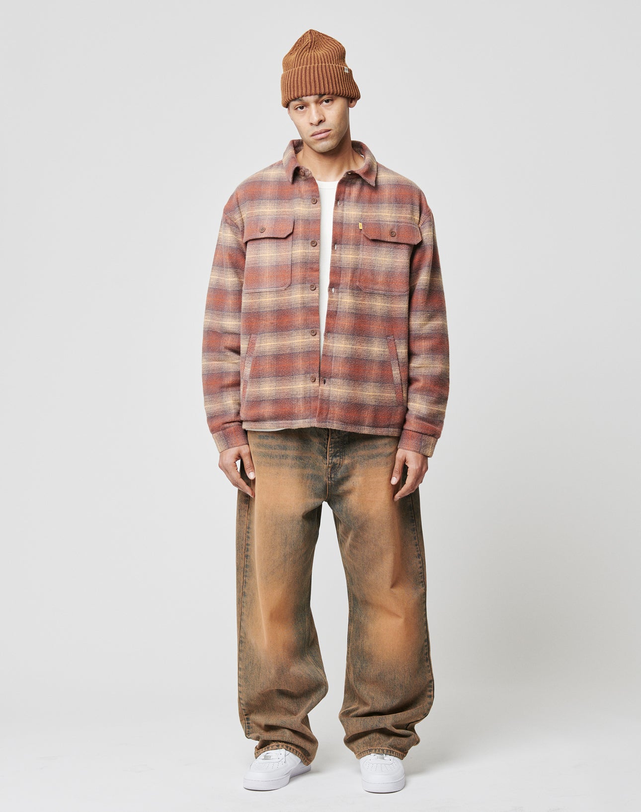 A man wears the LIVE FAST DIE YOUNG Vintage Padded Flannel in pink and brown plaid over a white tee, with loose-fit tan and brown vintage-wash jeans, white sneakers, and a brown beanie, standing against a white background.
