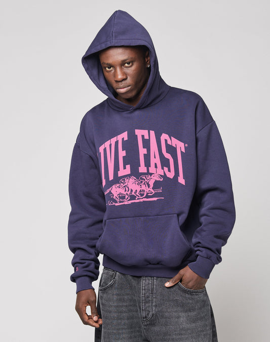 A person in the LIVE FAST DIE YOUNG Live Fast Stallion Hooded, an oversize dark purple hoodie aus Baumwolle with bold pink LIVE FAST frontprint above racing horses, pairs it with dark gray jeans against a plain light background.
