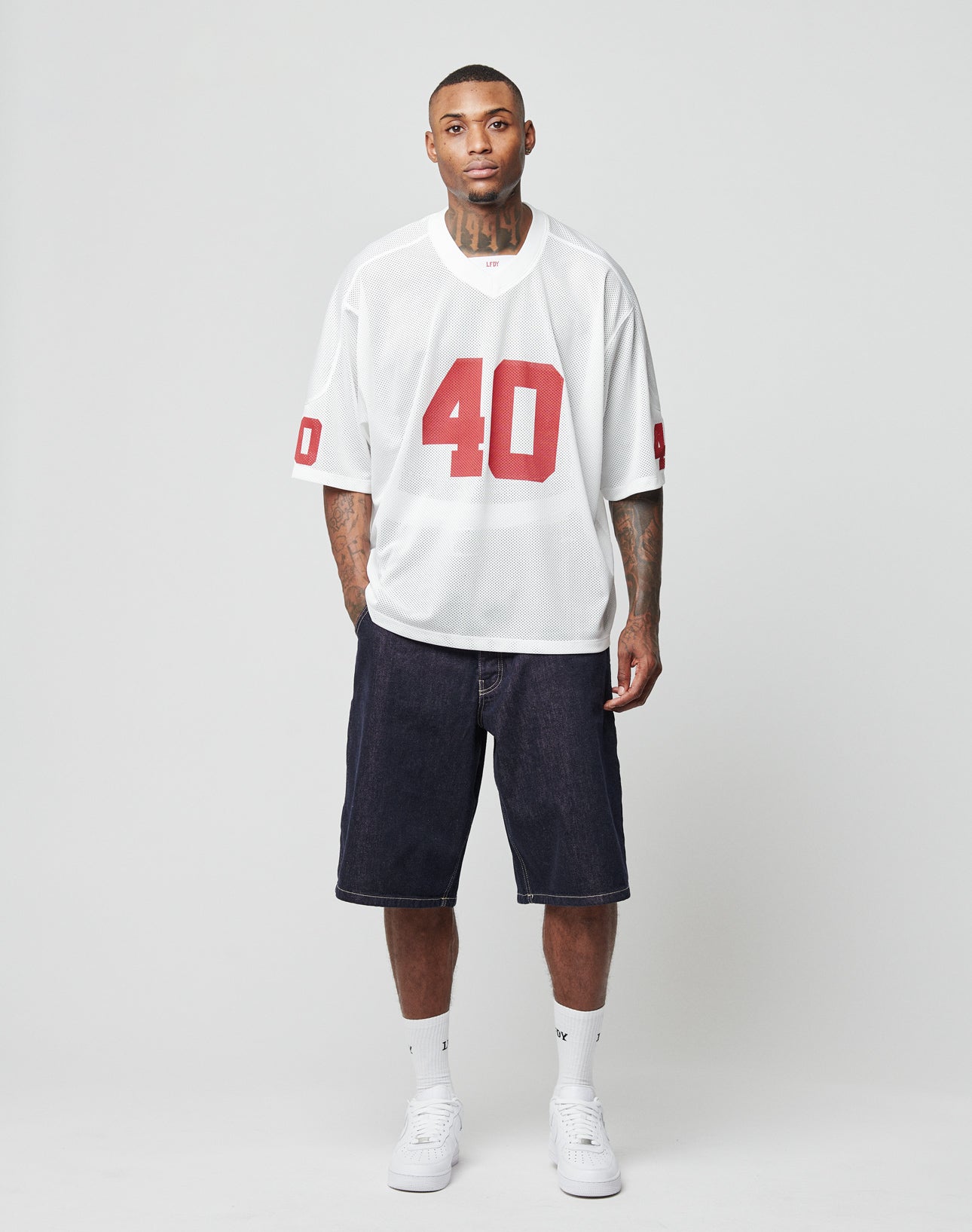 A man stands against a plain background wearing the LFDY Mesh Jersey by LIVE FAST DIE YOUNG, a white football jersey with red number 40, V-Ausschnitt, and Oversize Fit, styled with dark denim shorts, white socks, and white sneakers.