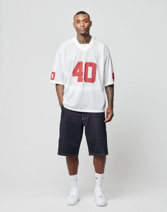 A man stands against a plain background wearing the LFDY Mesh Jersey by LIVE FAST DIE YOUNG, a white football jersey with red number 40, V-Ausschnitt, and Oversize Fit, styled with dark denim shorts, white socks, and white sneakers.