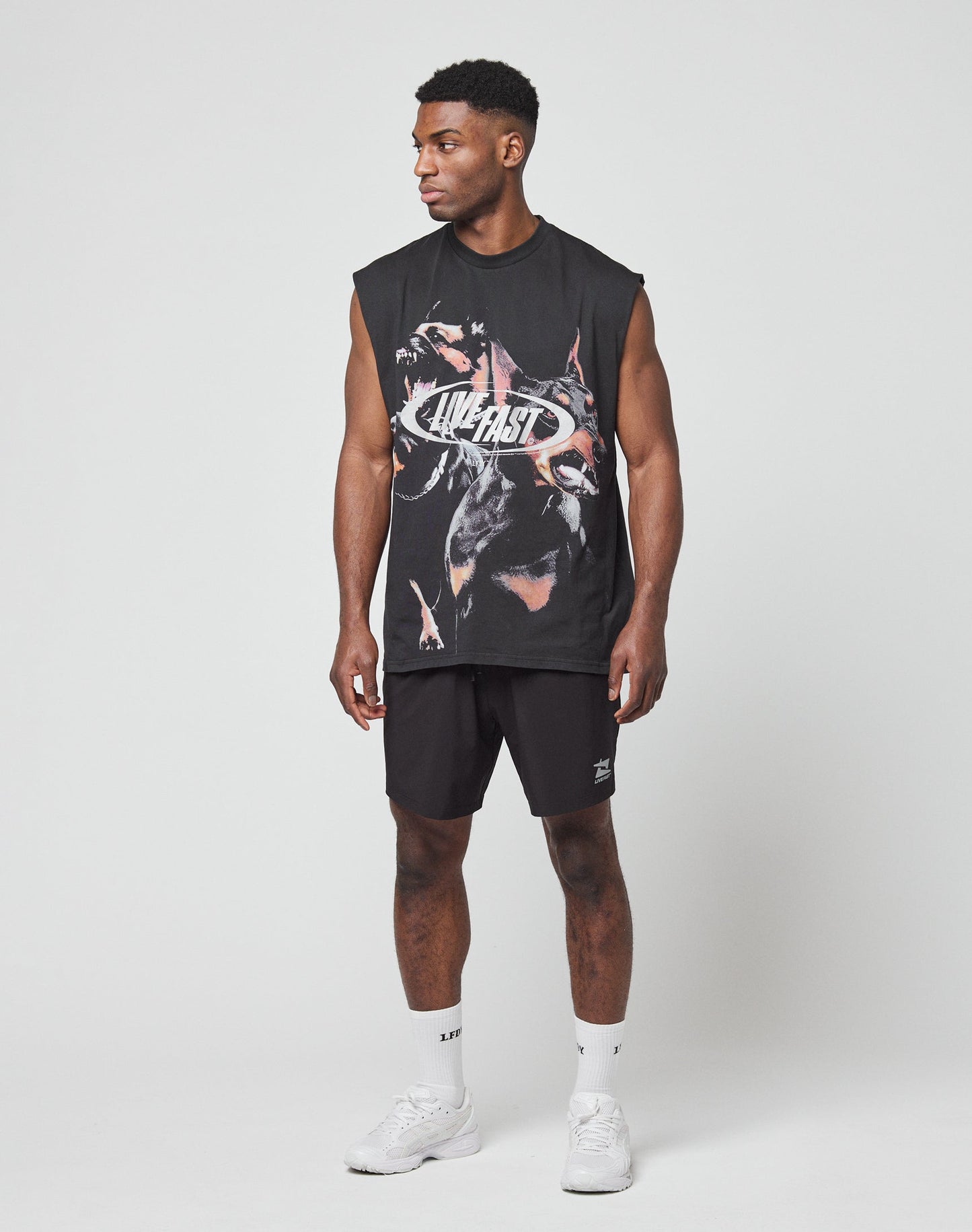 A man stands against a plain white background, wearing a Regular Fit black graphic sleeveless shirt with “LIVE FAST” text, LIVE FAST DIE YOUNG Live Fast Performance Shorts, white socks, and white sneakers. He looks slightly left with arms relaxed.