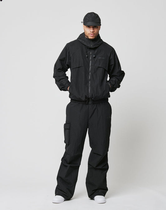 Against a plain white background, a person wears an oversized black zip-up jacket and LIVE FAST DIE YOUNG Live Fast Nylon Pants with an elastic waistband, paired with white sneakers. Their hands are in their jacket pockets.
