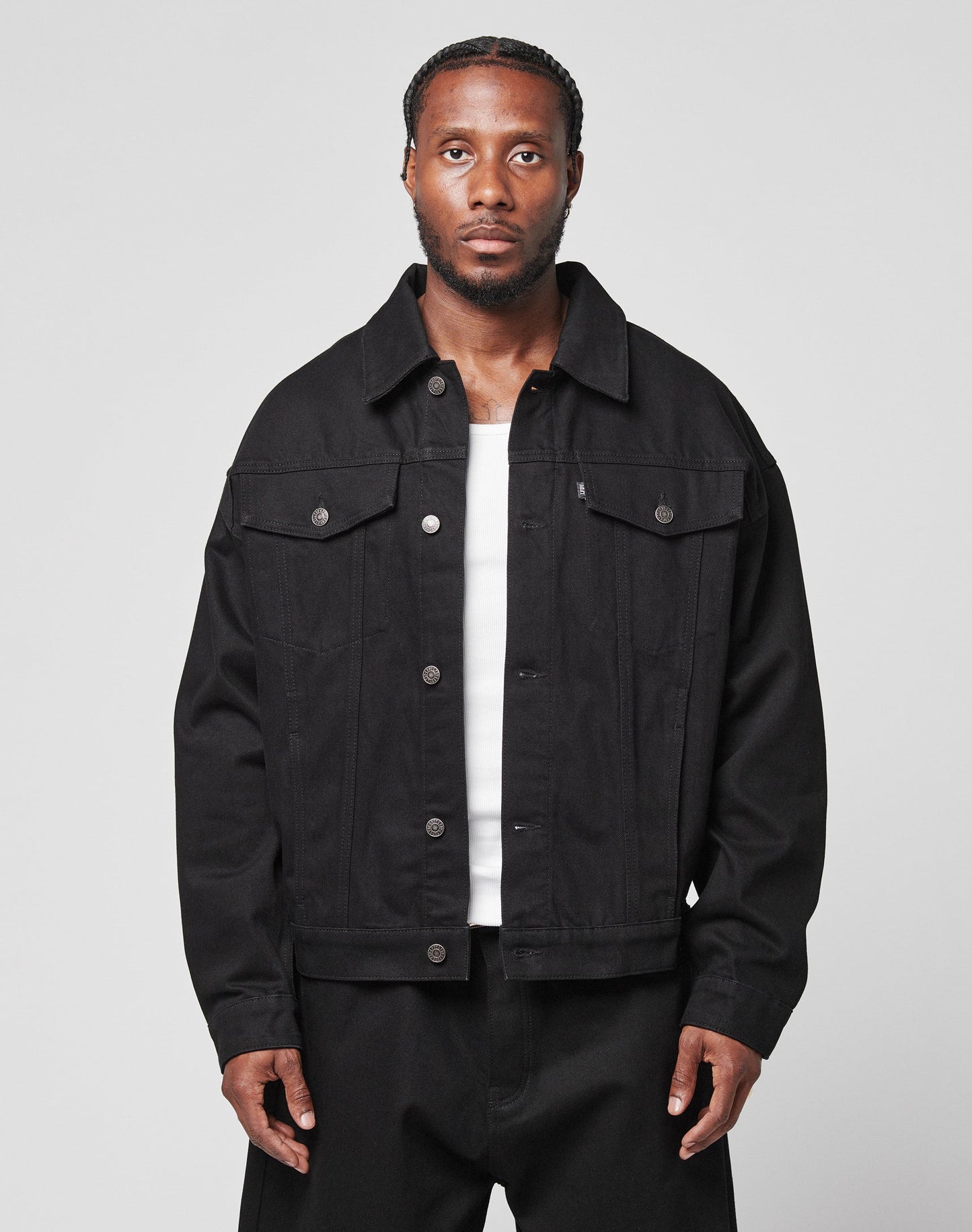 A man with braided hair wears an oversize fit LFDY Denim Jacket by LIVE FAST DIE YOUNG, made from 100% cotton, over a white shirt and black pants. He stands against a plain light gray background, looking directly at the camera with a neutral expression. | model