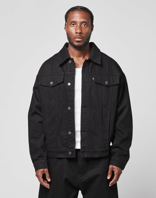 A man with braided hair wears an oversize fit LFDY Denim Jacket by LIVE FAST DIE YOUNG, made from 100% cotton, over a white shirt and black pants. He stands against a plain light gray background, looking directly at the camera with a neutral expression. | model