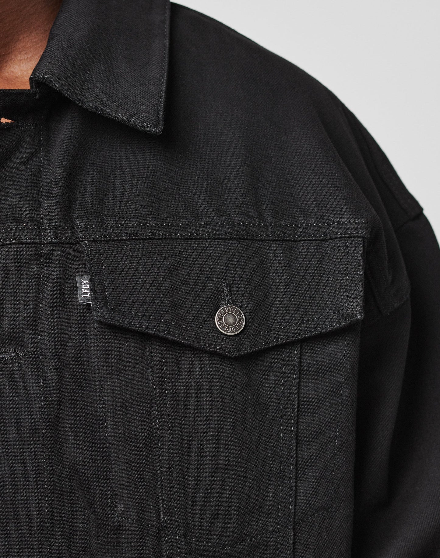 Close-up of the upper part of the LFDY Denim Jacket by LIVE FAST DIE YOUNG, made from 100% cotton with a metal button, small fabric tag on the chest pocket, pointed collar, and visible stitching.
