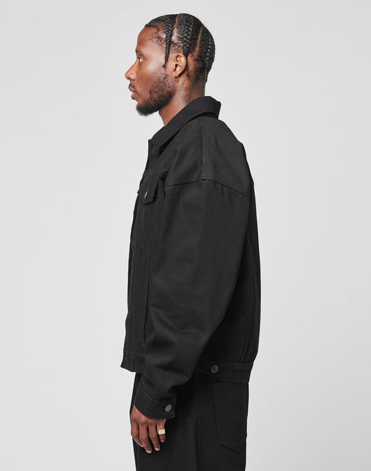 A man with braided hair, wearing the LFDY Denim Jacket by LIVE FAST DIE YOUNG and oversized black pants, stands in profile against a plain white background.