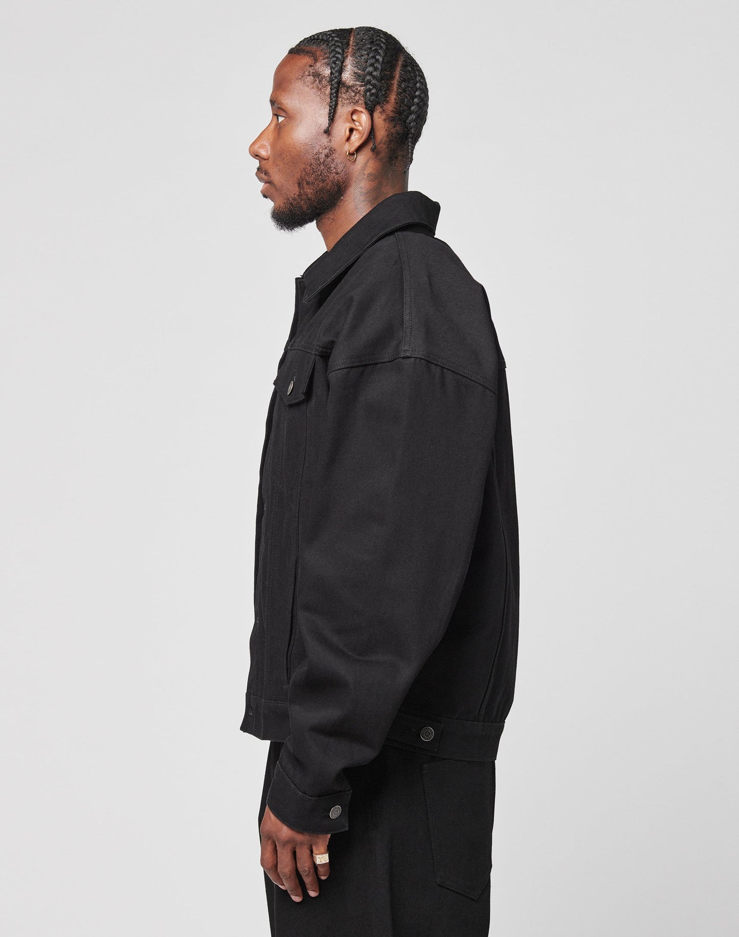 A man with braided hair, wearing the LFDY Denim Jacket by LIVE FAST DIE YOUNG and oversized black pants, stands in profile against a plain white background.