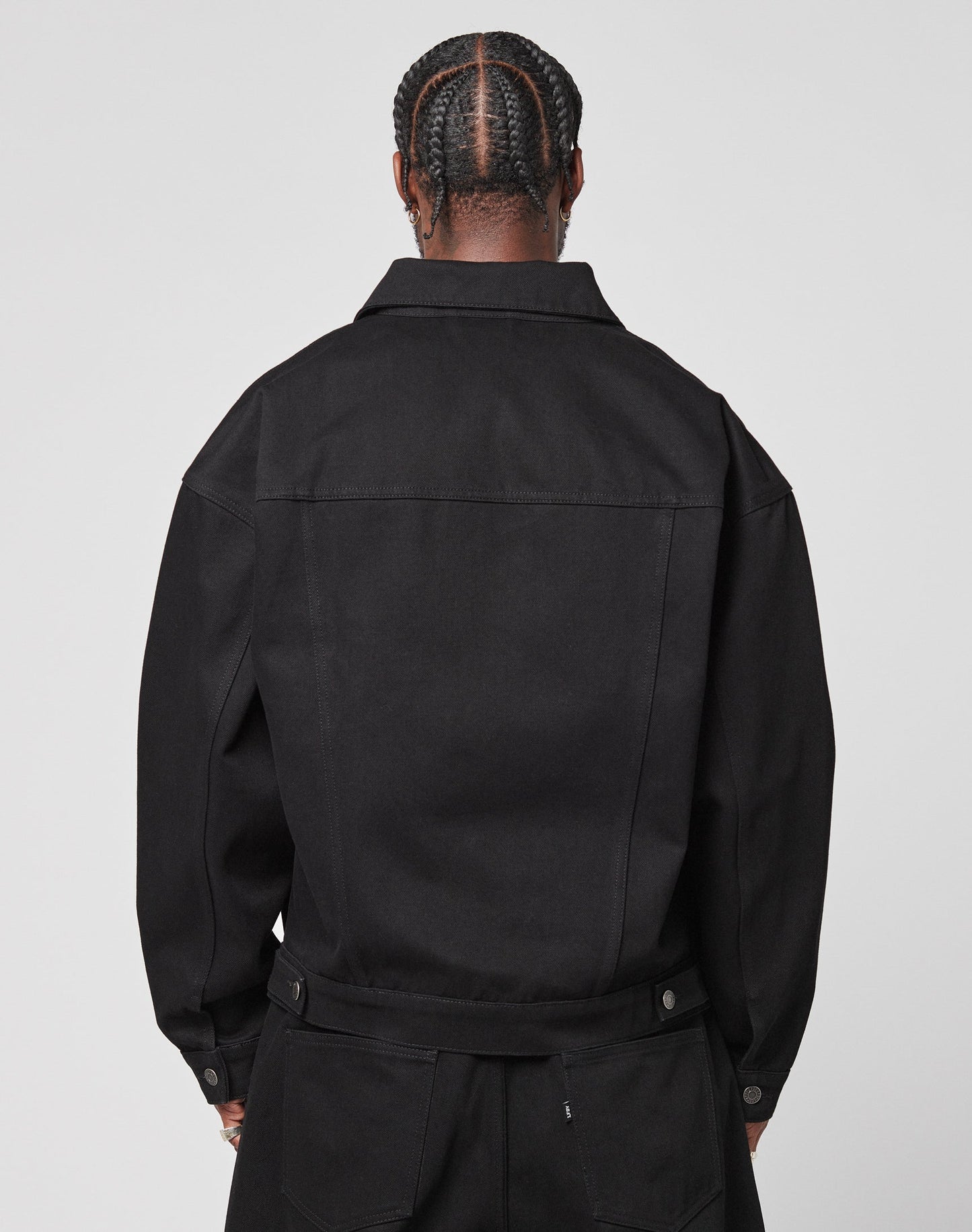 A person with neatly braided hair is shown from behind, wearing an oversize fit black LFDY Denim Jacket by LIVE FAST DIE YOUNG and matching black pants against a plain light background.
