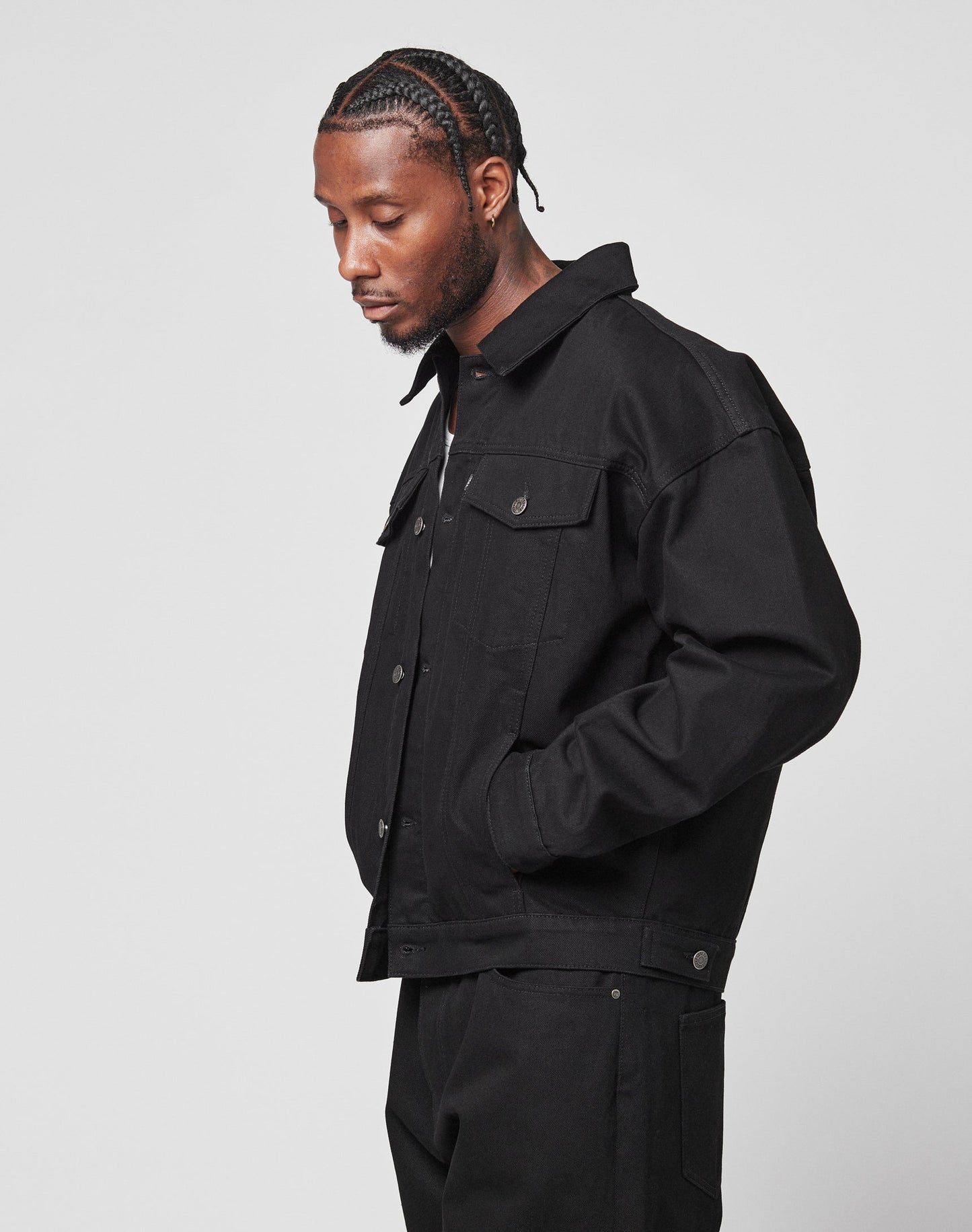 A man with braided hair wears the LFDY Denim Jacket by LIVE FAST DIE YOUNG and matching black pants, standing against a plain white background with his head slightly bowed and one hand in his pocket.