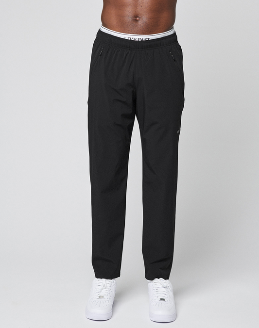 A person models LFDY Performance Pants by LIVE FAST DIE YOUNG—black joggers with zippered pockets, an elastic waistband, and an oversize fit—paired with white sneakers against a plain light gray background.