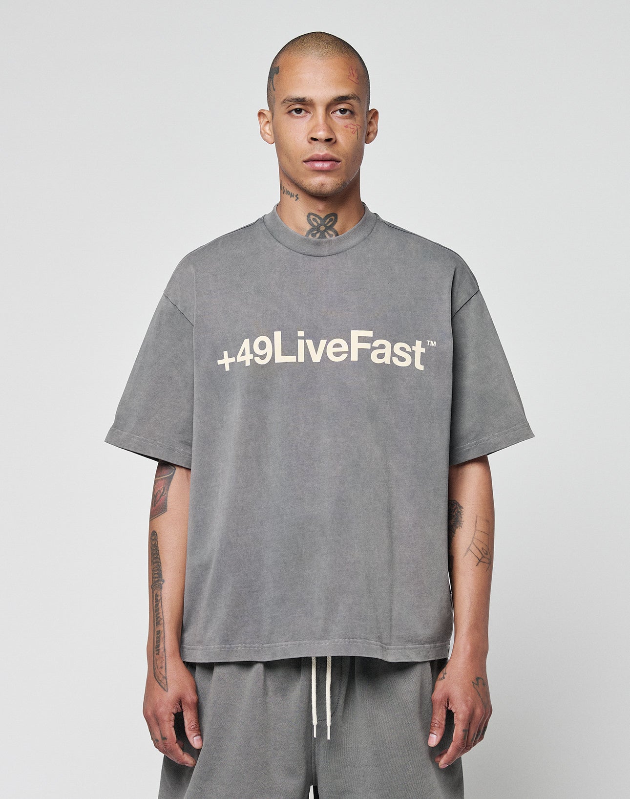 A tattooed, shaved-head individual wears the oversize-fit Basic +49 Tee by LIVE FAST DIE YOUNG, in gray 100% Baumwolle with a bold +49LiveFast front print, against a plain light background.