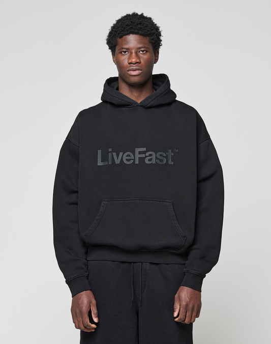 A person wearing the LIVE FAST DIE YOUNG Basic Livefast Hooded in black, featuring a bold "LiveFast" front print and an oversize fit, pairs it with black pants against a plain light gray background. | model