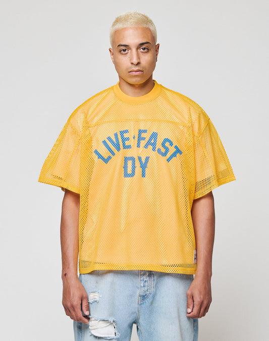 A person with short bleached blonde hair wears the LFDY Boxy Jersey by LIVE FAST DIE YOUNG, a yellow mesh T-shirt with blue LIVE FAST D(Y) lettering, paired with light blue ripped jeans against a plain background.| model