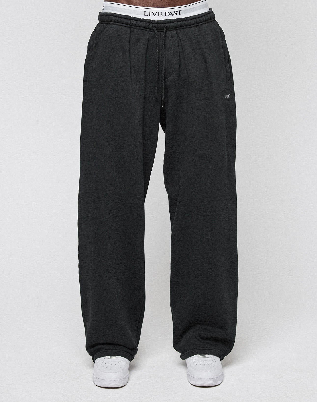 A person in LIVE FAST DIE YOUNG Basic Wide Jogger (black), white sneakers, and LIVE FAST waistband underwear stands against a plain light gray background.