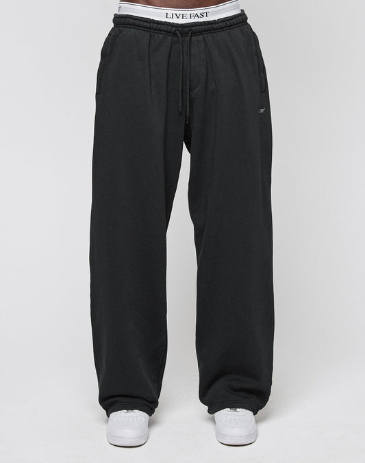A person in LIVE FAST DIE YOUNG Basic Wide Jogger (black), white sneakers, and LIVE FAST waistband underwear stands against a plain light gray background.