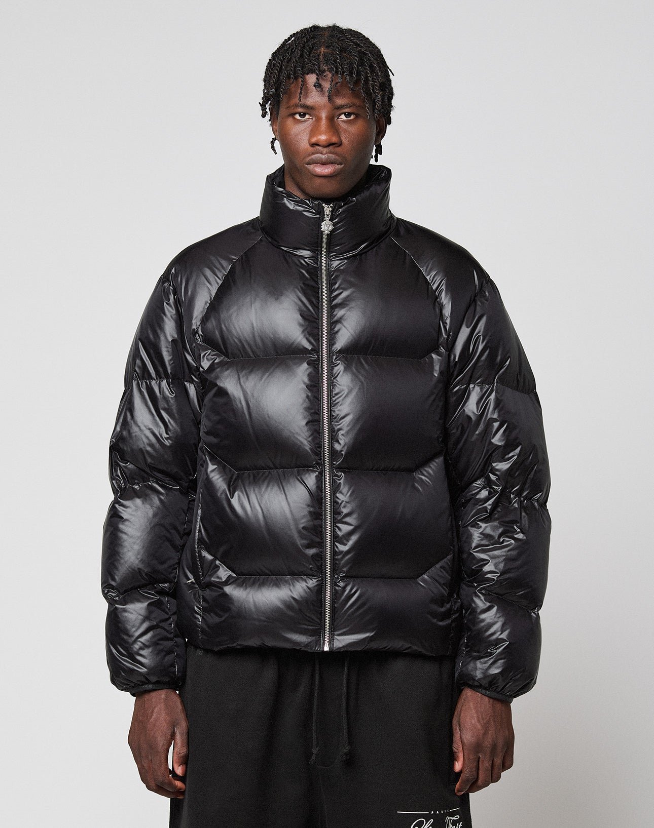 A person with short dreadlocks wears the shiny, wasserabweisend Live Fast Down Jacket by LIVE FAST DIE YOUNG, featuring a high collar, paired with black pants, standing against a plain light gray background. | model