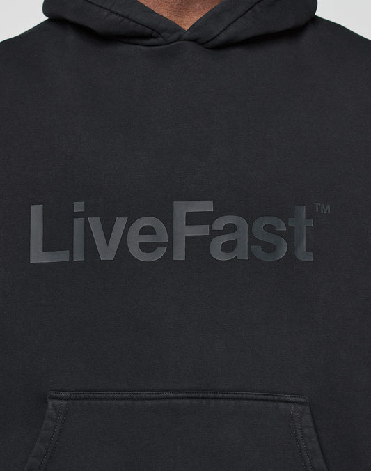 A close-up of the LIVE FAST DIE YOUNG Basic Livefast Hooded in black, featuring an Oversize Fit and bold LiveFast™ Frontprint across the chest, with partial views of the hood and front pocket.