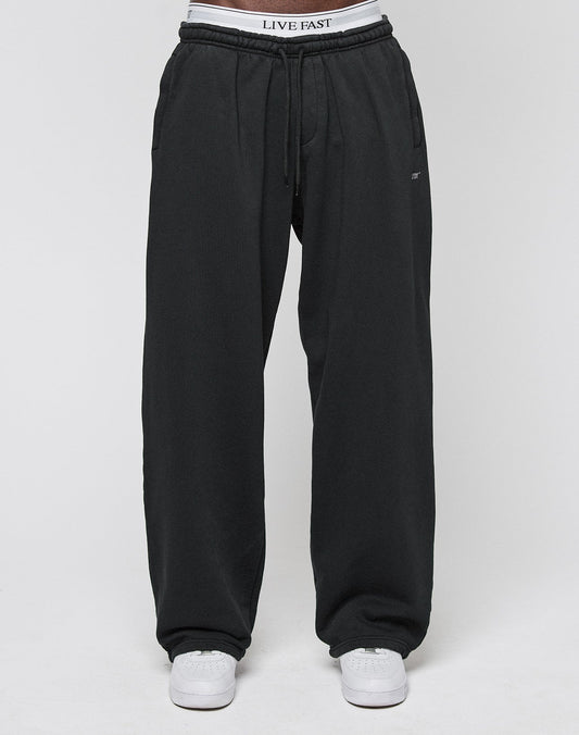 A person wearing the Basic Wide Jogger in black by LIVE FAST DIE YOUNG, featuring an elastic waistband with LIVE FAST text, paired with white sneakers, is standing against a plain white background.