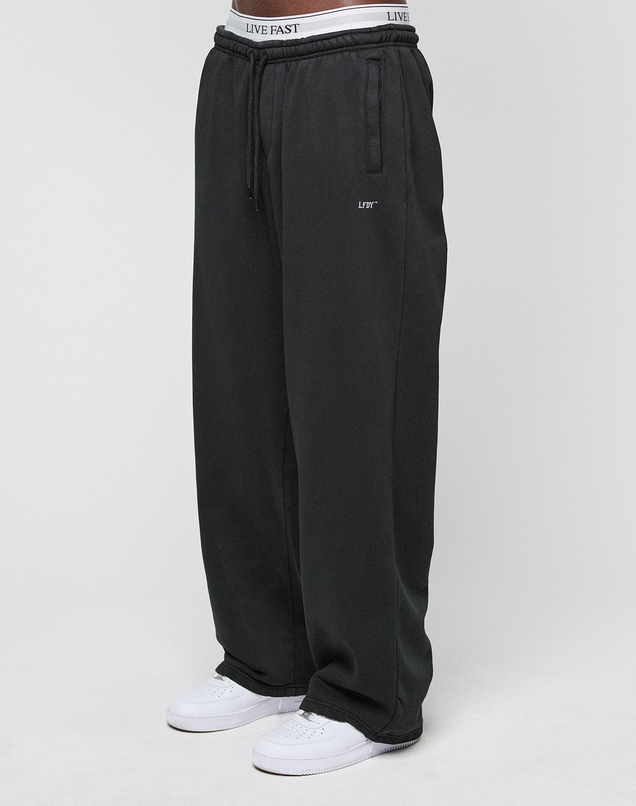 A person wears the LIVE FAST DIE YOUNG Basic Wide Jogger in black with a drawstring waist, white sneakers, and visible LIVE FAST branded underwear. The background is plain and light-colored. | model