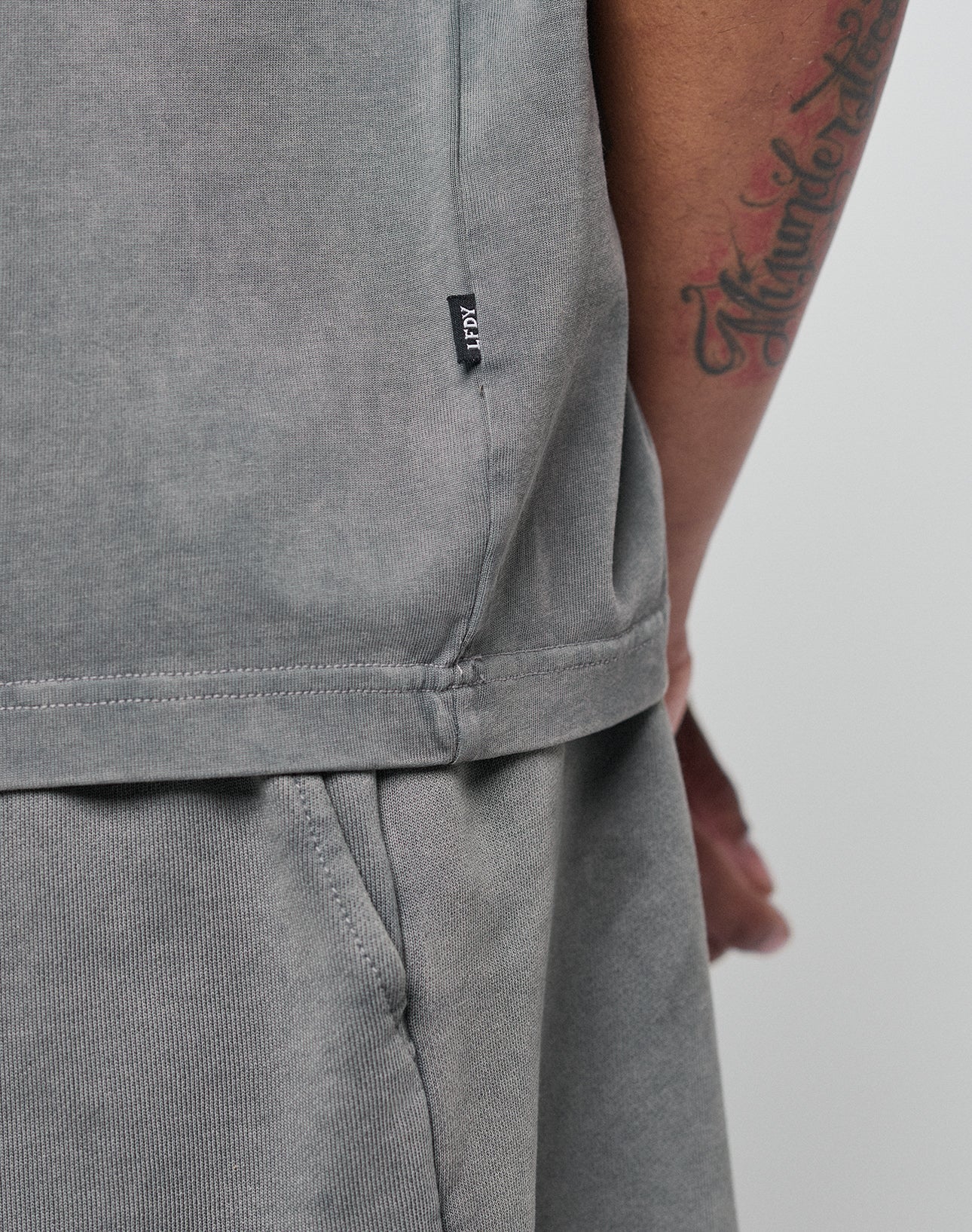 A close-up of a person’s arm and side wearing the LIVE FAST DIE YOUNG Basic +49 Tee in grey. Made from 100% Baumwolle with an oversize fit, it features a small black ICON tag and shows part of a tattoo on the forearm.