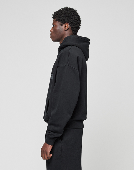 A person with short curly hair, seen in profile facing left, wears a black LIVE FAST DIE YOUNG Basic Livefast Hooded and black pants against a plain light gray background.